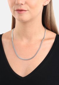 Srebrny łańcuch naszyjnik o płaskim, wzorze z zaprasowanymi ogniwami. Posiada gładkie, polerowane wykończenie metalowe, noszony na czarnej bluzce z dekoltem w serek.