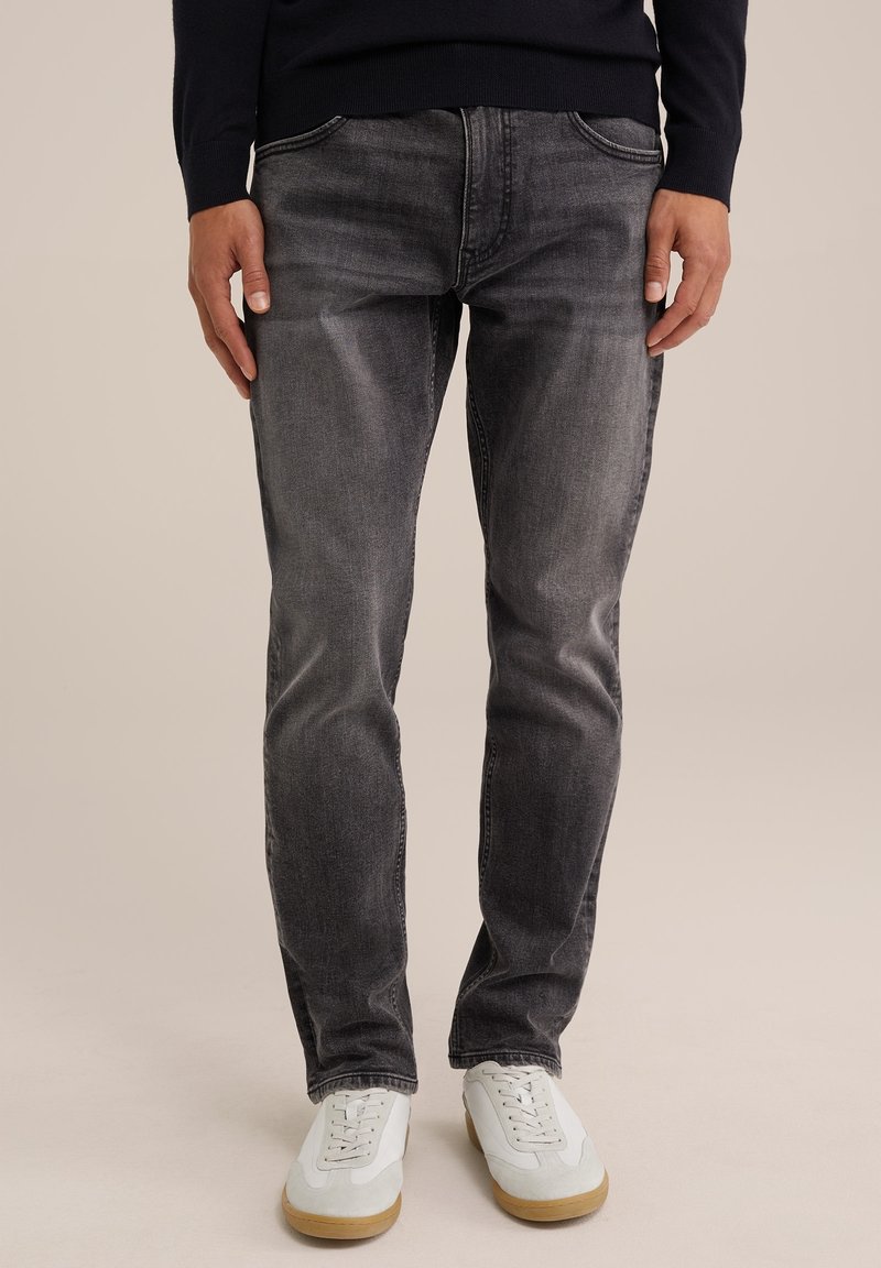 Man som bär urblekta svarta jeans och vita sneakers, står med händerna avslappnat vid sidorna mot en neutral bakgrund.