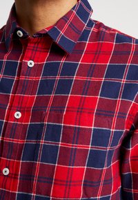 Czerwona i granatowa koszula w kratę zapinana na guziki, z kołnierzykiem, białymi guzikami oraz kieszenią na piersi z kontrastującym wzorem kraty.
