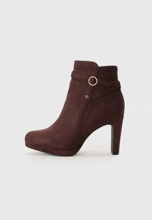 Bottine en suede marron avec un bout arrondi, un talon haut et une sangle décorative avec un détail en forme d'anneau doré. Texture lisse et finition brillante.