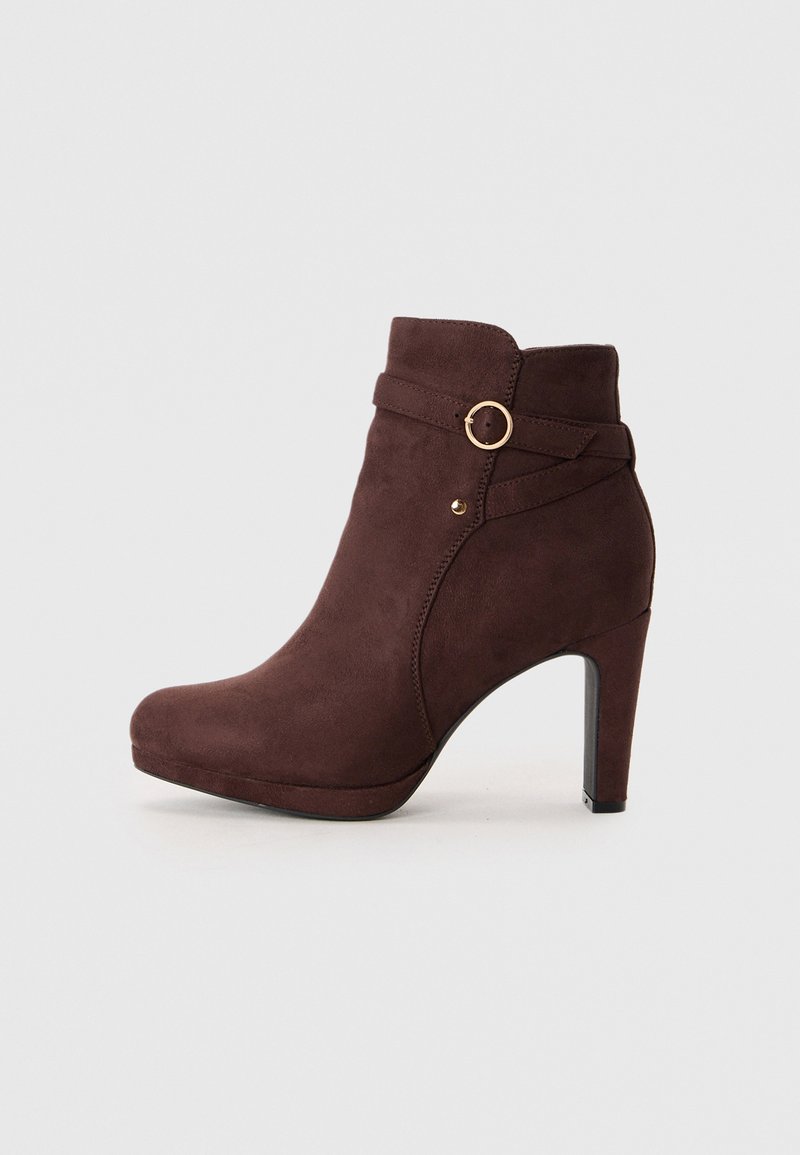 Bottine en suede marron avec un bout arrondi, un talon haut et une sangle décorative avec un détail en forme d'anneau doré. Texture lisse et finition brillante.