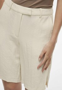 Shorts in lino beige chiaro con superficie texture, tasche laterali e taglio ispirato alla natura, caratterizzati da una vita alta e una parte frontale piatta.