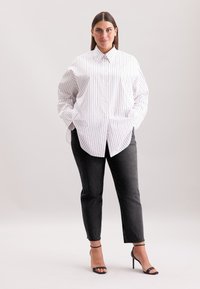 Donna in piedi con le mani nelle tasche, che indossa una camicia bianca a righe larghe, jeans neri e sandali neri con tacco alto, su uno sfondo neutro.