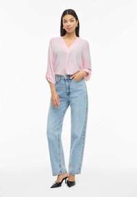 Femme debout portant un chemisier à col en V rose clair, un jean taille haute bleu clair et des chaussures à talons pointues noires.