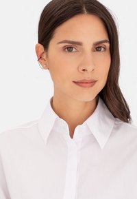 Weißes, knopfbares Hemd mit strukturiertem Kragen, das eine glatte Textur aufweist. Das Modell trägt minimale Accessoires und hat glattes Haar.