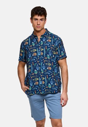 Hombre con camisa azul de manga corta con botones y patrones de peces coloridos, y pantalones cortos azul claro, de pie con una mano en el bolsillo.