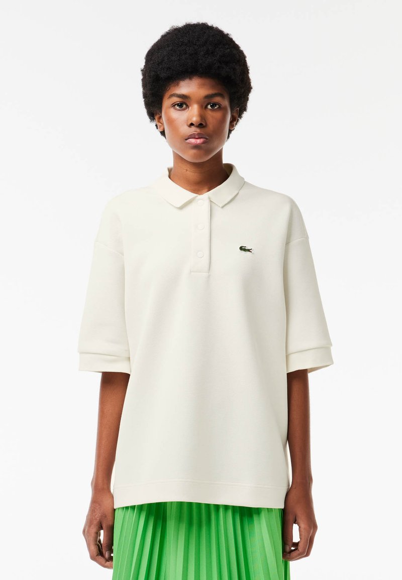Polo bianco a maniche corte con tessuto strutturato, caratterizzato da un piccolo logo verde sul petto, abbinato a una gonna verde plissettata.