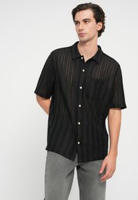 Trumpas rankovės juodas marškinėlių užsegimas su tekstūruotu, raižytu dizainu ir viena krūtinės kišene. Yra šviesių spalvų mygtukai ir apykaklė.