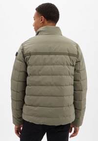 Man in olijfgroene, gequiltte pufferjack met hoge kraag, gezien vanaf de achterkant, staand tegen een witte achtergrond.