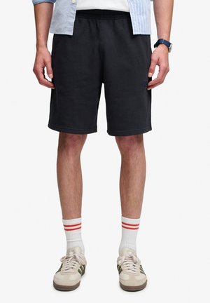 Uomo che indossa pantaloncini neri casual, calzini bianchi con due strisce rosse, sneakers beige con strisce verdi, orologio blu e camicia a righe azzurro chiaro.