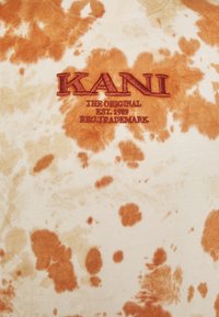 Tkanina z bavlny s batikováním v oranžových a béžových vzorech, s červeným výšivkovým nápisem "KANI THE ORIGINAL EST. 1989 REG. TRADEMARK".