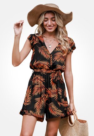 Mujer con mono corto negro con estampado de hojas de palma naranja, que lleva un sombrero de paja de ala ancha y sostiene un bolso de paja tejido, sonriendo con los ojos cerrados.