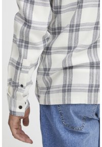 White plaid shirt with black lines, buttoned cuffs, set against blue denim jeans. Smooth fabric with a relaxed fit, textured pattern detail.