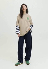 T-shirt oversized bege com gráfico verde, sobreposto a mangas compridas listradas em azul-marinho. Combinado com calças largas azul-marinho e ténis verdes.