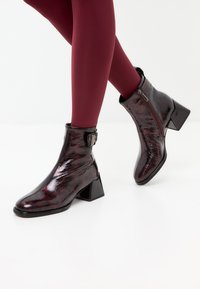 Bottines bordeaux à finition brillante, avec des talons carrés, un détail de fermeture éclair et un accent de boucle sur le côté. Associées à des collants marron.