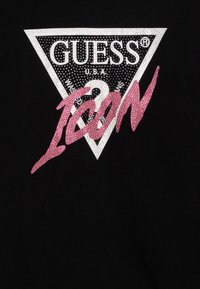 Black fabric with a silver studded triangle logo reading GUESS U.S.A. and a large pink studded script "ICON" overlapping the triangle.
