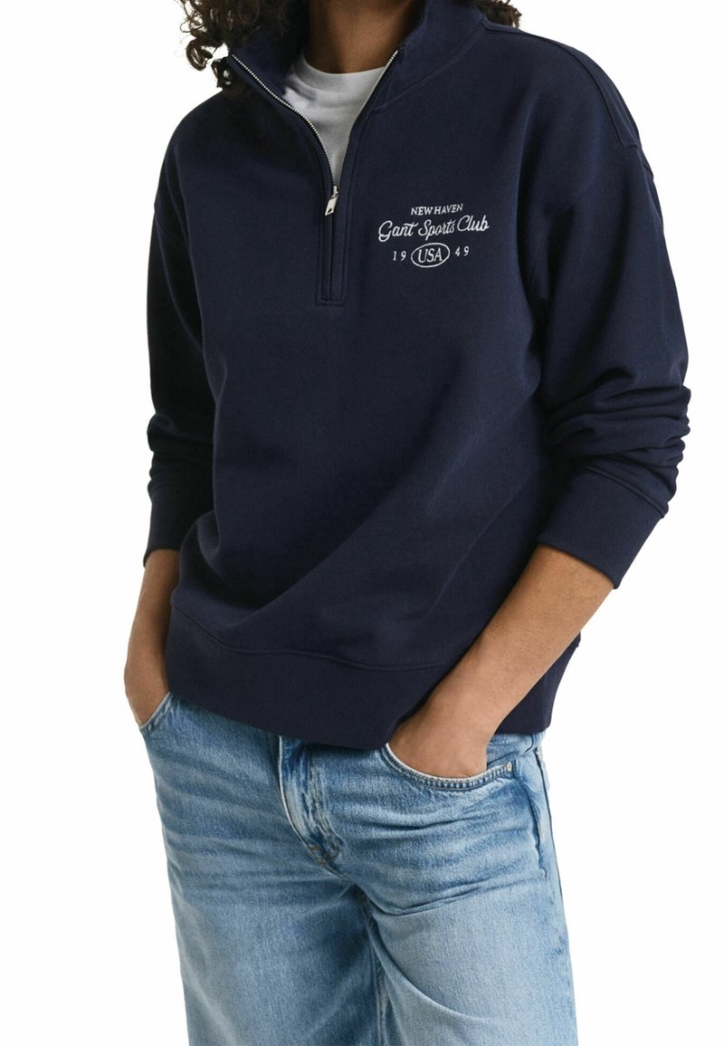 Persoon draagt een marineblauw kwart-ritssluiting sweatshirt met de tekst "New Haven Gant Sports Club 1949 USA", gecombineerd met lichtblauwe jeans.