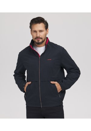 Mężczyzna w granatowej kurtce na zamek, pod którą ma białą koszulę, z rękami w kieszeniach, na tle jednolitego jasnoszarego tła.