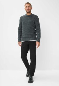 Maglione grigio scuro lavorato a maglia con scollo rotondo e polsini a costine, abbinato a pantaloni neri e scarpe nere lucide, in piedi su uno sfondo bianco.