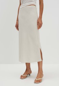 Langer, beigefarbener Rock aus leichtem Stoff mit glatter Textur. Verfügt über einen Seitenschlitz und ein gerades Schnittdesign. Kombiniert mit hautfarbenen Sandalen.