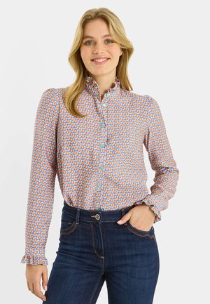 Joven sonriente, vestida con una blusa abotonada de patrones coloridos, con cuello y puños con volantes, combinada con jeans azul oscuro.
