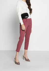Donna che indossa un maglione bianco a maglia, pantaloni cropped rosa antico, tacchi alti con stampa leopardata e porta una piccola borsa a tracolla nera.