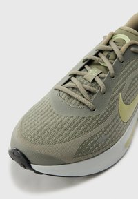 Green athletic shoe with textured upper, contrasting beige accents, and a rounded toe. Features laces and a distinctive logo.