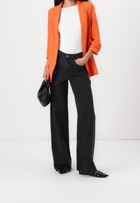 Blazer naranja sobre una blusa blanca, combinado con jeans de tiro alto de pierna ancha negros. Bolso negro y zapatos planos puntiagudos con detalles de ojales.