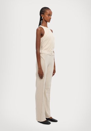 Top senza maniche beige abbinato a pantaloni larghi beige in tessuto testurizzato, con dettagli di cucitura sottili e scarpe nere piatte.