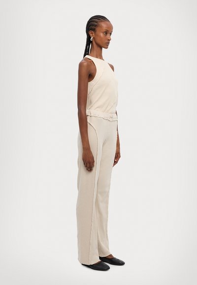 Haut sans manches beige associé à un pantalon large beige en tissu texturé, avec des détails de couture subtils, et des chaussures noires plates.