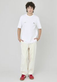 T-shirt blanche avec le texte "Meilleur de sa classe", associée à un pantalon crème et des baskets rouges avec des rayures blanches. Coupe modérée, style décontracté.