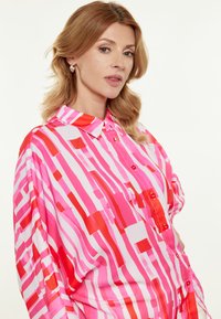 Rosa gestreifte Bluse mit unterschiedlich breiten roten und hellrosa Linien, einem Kragen und roten Knöpfen auf der Vorderseite. Glattes Material mit lockerer Passform.