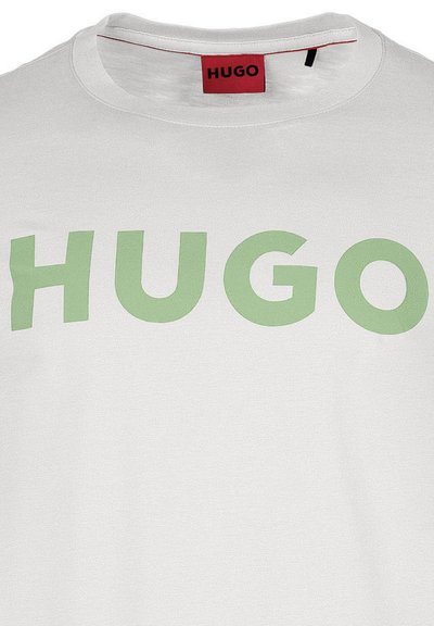 Бяла памучна тениска с големи, смели зелени букви "HUGO" отпред, с червен бранд етикет, видим на врата.