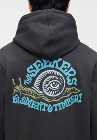 Personne portant un sweat à capuche sombre avec un grand dessin dans le dos représentant un escargot avec un œil sur sa coquille et un texte bleu indiquant "THE SEEKERS ELEMENT & TIMBER !"