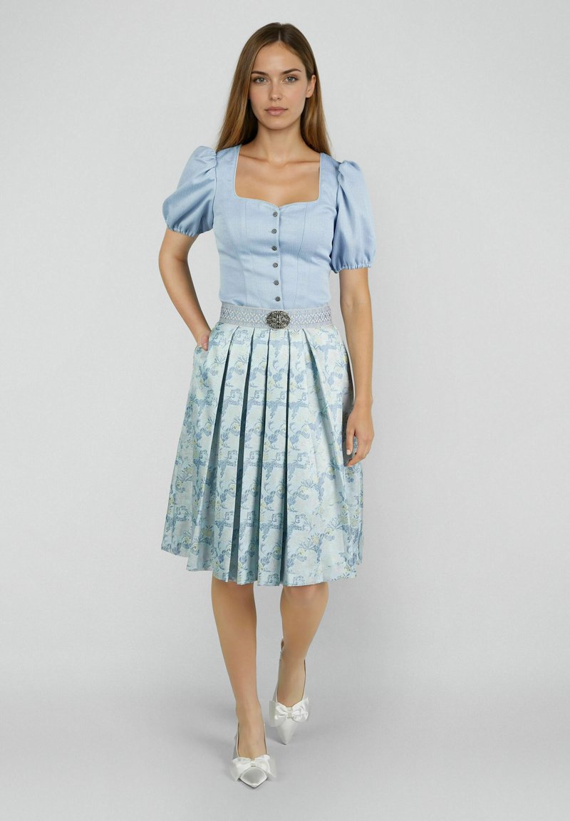 Frau in einem hellblauen traditionellen Kleid mit Puffärmeln, floralem Faltenrock, dekorativem Gürtel und weißen spitzen Schuhen mit Schleifen.