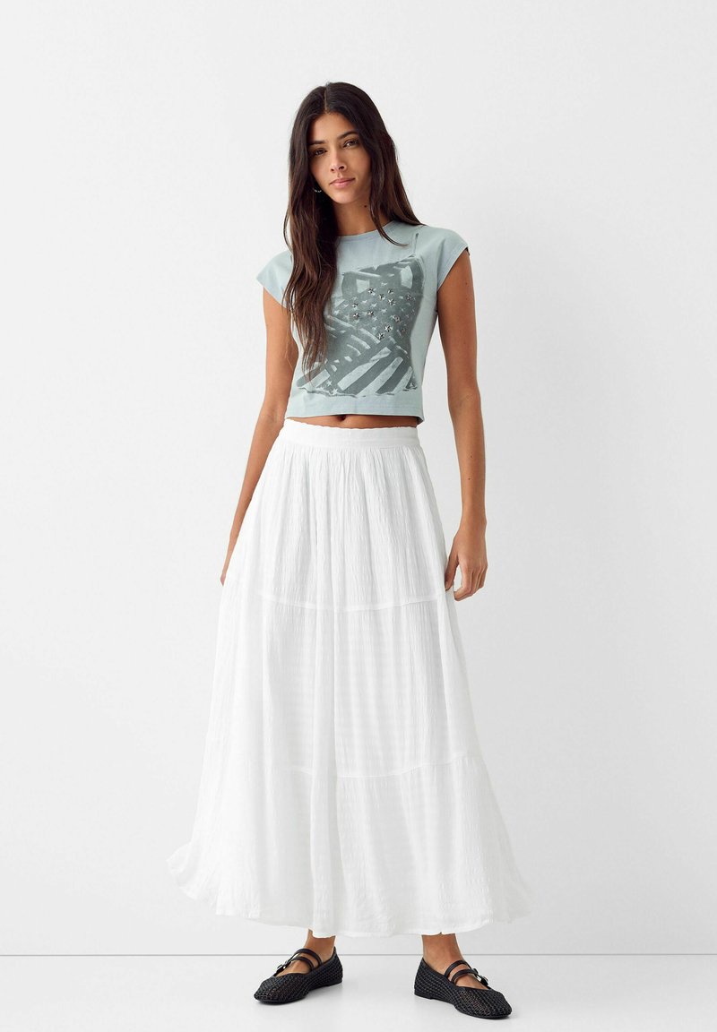 Lichtblauwe cropped t-shirt met grafische print, witte gelaagde maxi rok en zwarte geweven schoenen. Zachte texturen en casual ontwerp.