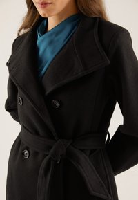 Vrouw met een zwart mantel met dubbele knopen en ceintuur, gedragen over een teal satijnen blouse, getoond van hals tot heupen.