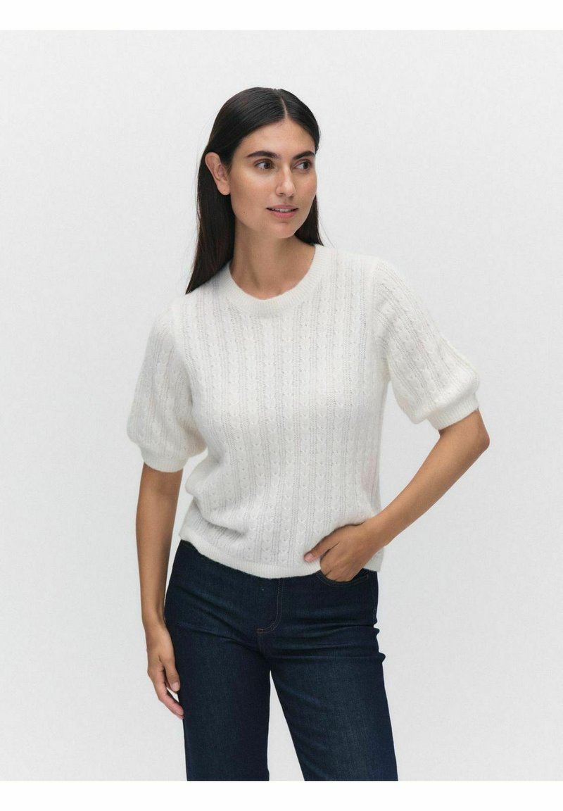 Vit stickad tröja med korta, voluminösa ärmar, som har ett strukturerat mönster och ribbad fåll. Bärs med mörka jeans.