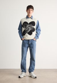 Mladý muž stojící s rukama v kapsách, oblečený v modrých džínách, denimové košili, krémovém svetr vestě s černým geometrickým květinovým vzorem a bílých teniskách.