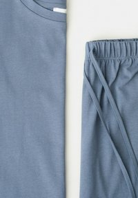 Camicia di cotone blu piegata con bordo bianco sul colletto accanto a pantaloni blu con coulisse abbinati, su sfondo bianco.