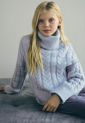 Ragazza bionda con occhi azzurri che indossa un maglione a collo alto lavorato a trecce di colore viola chiaro, seduta su una superficie grigia contro uno sfondo semplice.