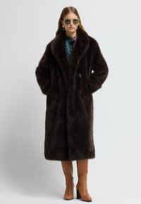 Abrigo de pelo sintético marrón con un amplio cuello, dos bolsillos frontales y cierre de botón. Llevado sobre una blusa de estampado y combinado con botas de tacón en color marrón claro.