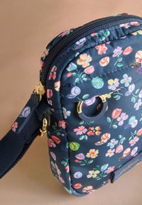 Liten marinblå tygväska med korsrem, färgglatt blommönster, guldfärgad dragkedja och ringdetaljer samt justerbar marinblå rem mot en neutral bakgrund.