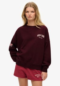 Sudadera burdeos con "NEW YORK" y "Superdry" bordados en blanco. Cuenta con un corte holgado, cuello redondo y un parche de la bandera americana en la manga.