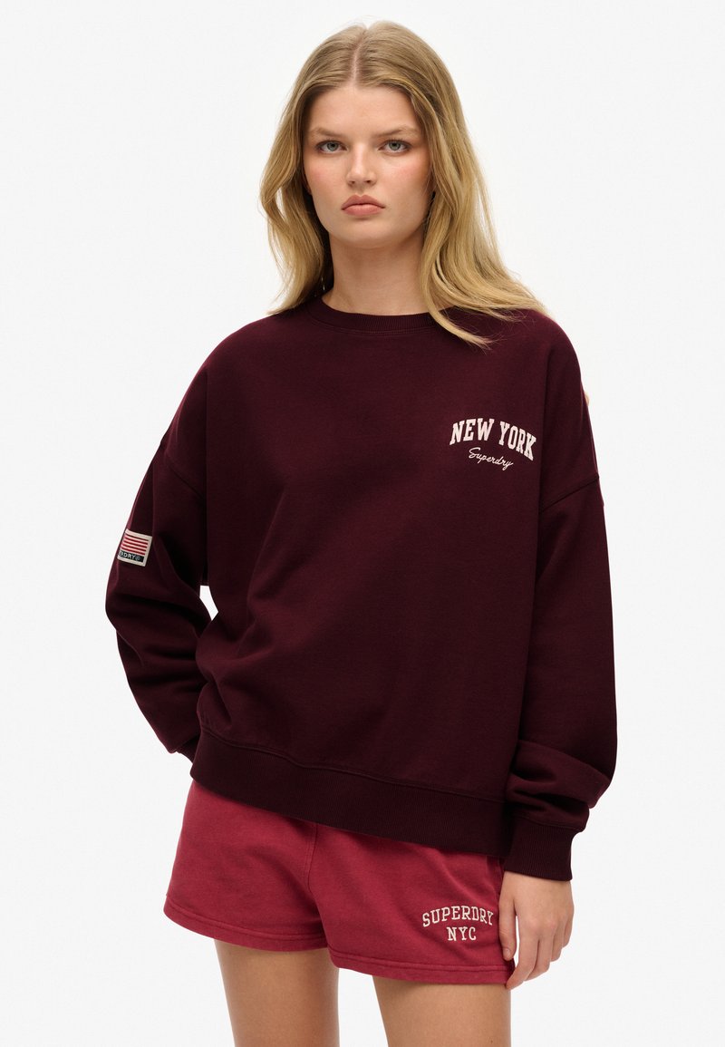 Sudadera burdeos con "NEW YORK" y "Superdry" bordados en blanco. Cuenta con un corte holgado, cuello redondo y un parche de la bandera americana en la manga.