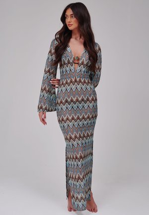Femme aux longs cheveux foncés portant une robe à manches longues et longue, à motif ondulé bleu, marron et beige, debout pieds nus sur un fond uni.