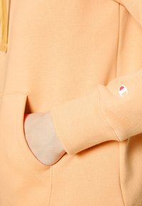 Peach-värinen huppari pehmeästä kankaasta, jossa kengurutasku ja resorit hihansuissa. Vasemmassa hihassa on pieni logodetalji.