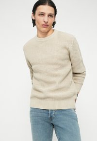Maglione beige a maglia con colletto rotondo, motivo testurizzato e orlo a costine, abbinato a jeans azzurri chiari.