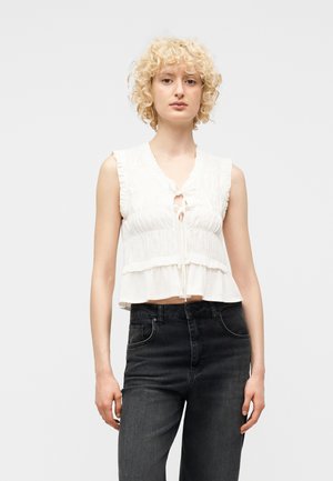 Osoba o blond kręconych włosach, ubrana w białą, bezrękawnikową bluzkę z falbanami i wiązaniem oraz czarne, wysokiego stanu dżinsy, stojąca na białym tle.