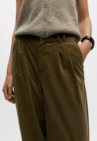 Khaki corduroy broeken met plooien, voorzien van zijzakken; gecombineerd met een gestructureerde beige gebreide vest en een zwarte armband.
